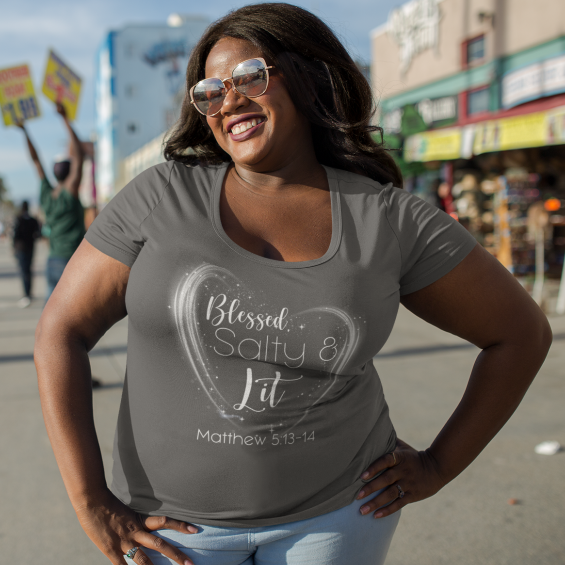 Woman wearing a gray t-shirt with text, standing on a street with palm trees and buildings in the background.