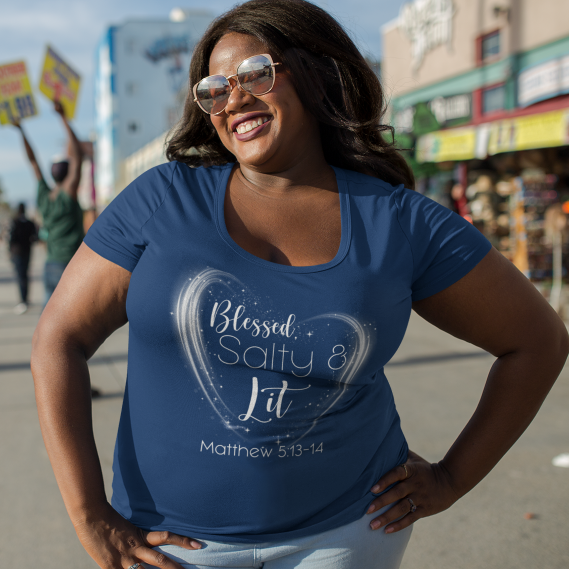 Woman wearing a blue t-shirt with text and graphics, standing on a street with palm trees and buildings in the background.