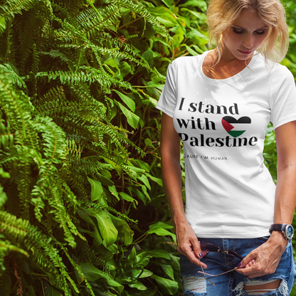 Woman wearing a white t-shirt with 'I stand with Palestine' text, standing in front of green foliage.